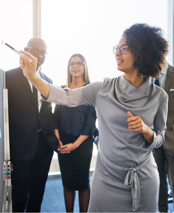 Woman stands in foreground writing on a white board while team members stand beside her watching