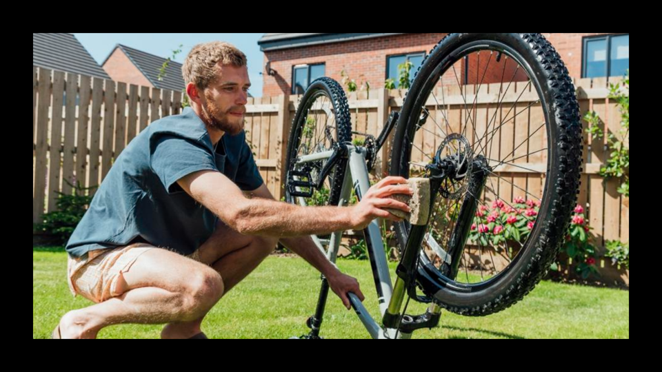 A man on a green lawn cleaning a bike that's tipped upside down with a sponge in the sun