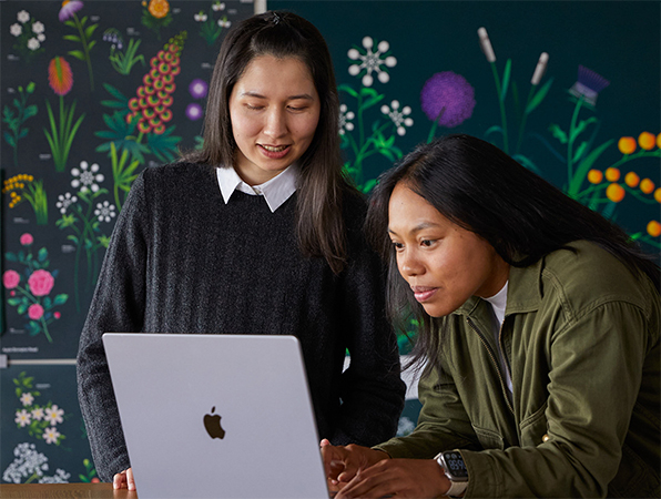 Zwei Apple Marketing Teammitglieder arbeiten gemeinsam an einem MacBook vor Postern, die ihr Team designt hat.