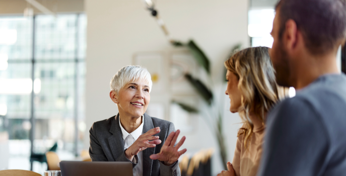 Professional female with short white hair sitting and talking to a young couple with their backsโ turned