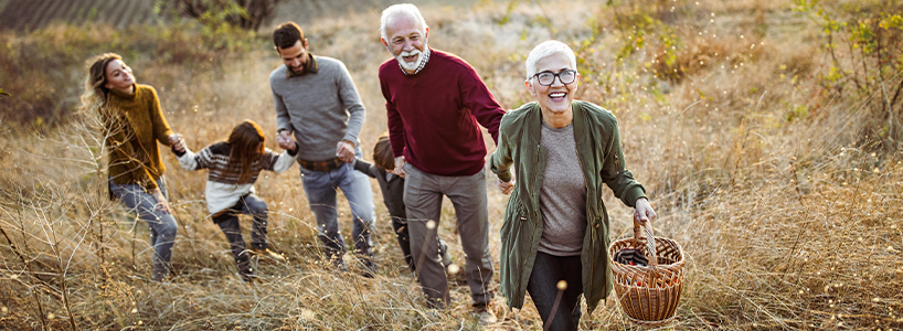 A mother and father, two children, and the children’s grandparents walk hand-in-hand in a field. The grandmother is carrying a basket.