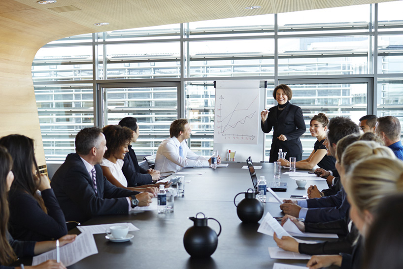 A woman standing in front of a graph on an easel presenting to an engaged group sitting around a boardroom table.